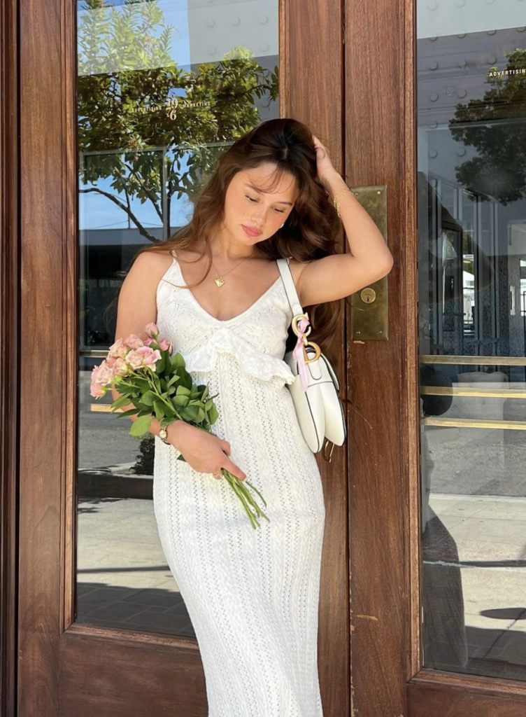 Woman in a white crocheted dress holding flowers by a door.